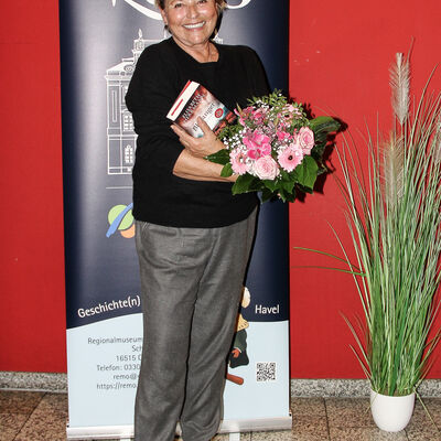 Elisabeth Herrmann mit ihrem Buch und einem Strau&szlig; Blumen in der Hand l&auml;chelt in die Kamera.