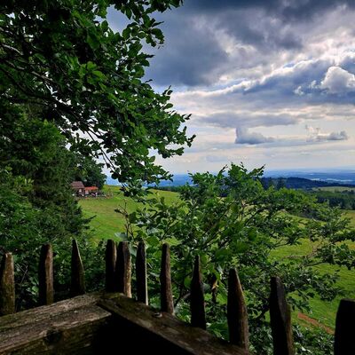 Das Bild zeigt eine gr&uuml;ne H&uuml;gellandschaft. Im Vordergrund sind B&auml;ume und ein rustikaler Holzzaun zu sehen, im Hintergrund ein blauer Himmel mit Wolken.