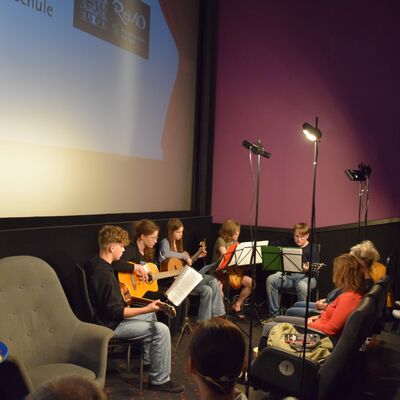 Jungs und M&auml;dchen sitzen mit Musikinstrumenten vor drei kleinen Scheinwerfern. Im Hintergrund ist eine gro&szlig;e Beamerleinwand zu sehen. Rechts im Bild sitzen M&auml;nner und Frauen im Publikum.