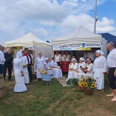 Wei&szlig;e Zelte mit polnisch-beschrifteten Schildern stehen auf einer Wiese. Im Vordergrund Frauen und M&auml;nner in wei&szlig;en Trachten.
