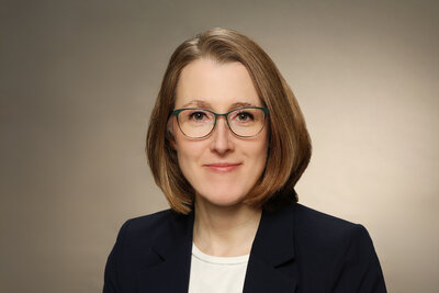 Eine Frau mit Brille mittleren Alters mit dunkelblonden, kinnlangen Haaren, schwarzem Sakko und wei&szlig;er Bluse. 