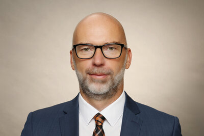 Ein Mann mit schwarzer Brille mittleren Alters ohne Haare, graumeliertem Vollbart mit dunkelblauem Sakko, dunkelgemusterter Krawatte und wei�em Hemd.