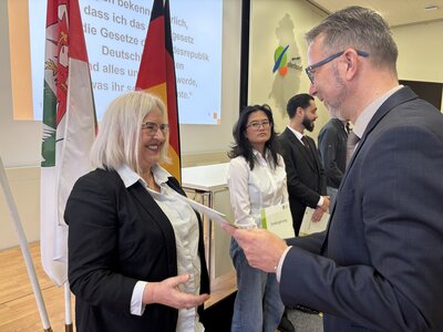 Ein Mann mittleren Alters im Anzug mit Brille (rechts im Bild) &uuml;berreicht einer Frau mit wei&szlig;blonden, schulterlangen Haaren und Brille ein Dokument. 