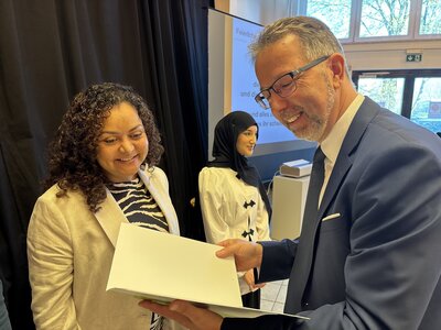 Ein Mann mittleren Alters mit Anzug, Hemd und Krawatte sch&uuml;ttelt einem kleinen Jungen mit Hemd und Weste die Hand. Im Hintergrund stehen zwei Frauen und ein weiterer Junge. 