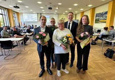Zwei M�nner und drei Frauen stehen in Zweierreihen in einem politischen Kreistagssaal und l�cheln in die Kamera. Zwei der Frauen und einer der M�nner halten Blumenstr�u�e in den H�nden.