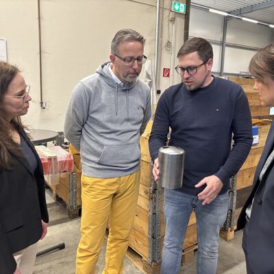 Zwei M&auml;nner und zwei Frauen stehen in einer Fabrikhalle. Ein Mann h&auml;lt einen metallischen Gegenstand in den H&auml;nden. Er scheint den anderen Personen daran etwas zu erkl&auml;ren.