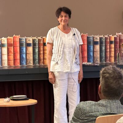 Eine &auml;ltere Frau mit kurzen, braunen Haaren steht vor einer braunen Wand. Direkt hinter ihr ein Regal mit zahlreichen B&uuml;chern.