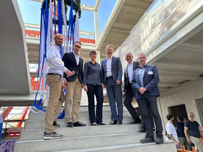 F&uuml;nf M&auml;nner und eine Frau stehen auf einem Rohbau einer Treppe auf einer Baustelle. &Uuml;ber ihnen h&auml;ngt eine Richtfestkrone mit blau-wei&szlig;en B&auml;ndern. 