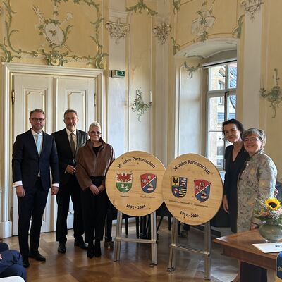 Drei Frauen und zwei M&auml;nner stehen in einem prunkvollen Schlosssaal neben zwei Holzschildern auf denen 35 Jahre Partnerschaft steht.